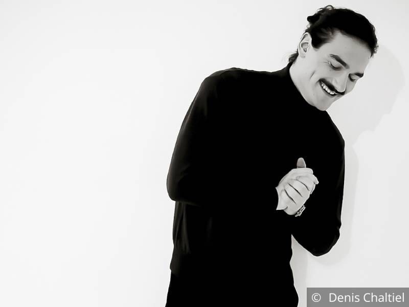 Photo en noir et blanc d'un jeune homme brun vêtu de noir qui pose les yeux fermé, souriant, les mains jointes.